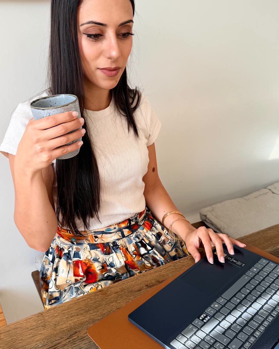 Adraa working at her table at home, a cup of tea in one hand and the other on her laptop.